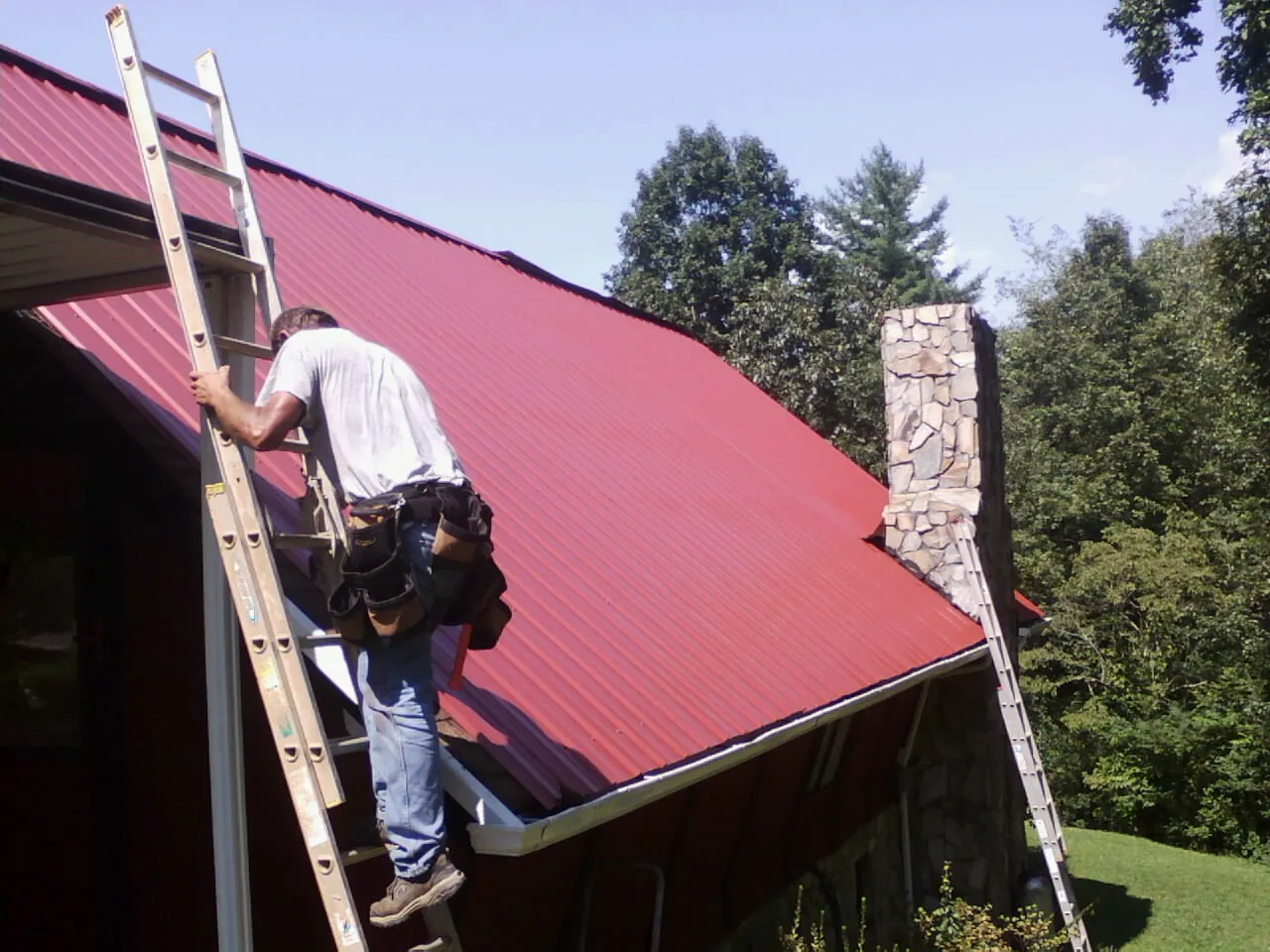Roofer on a ladder beside a red metal roof and stone chimney — Soffit & Fascia Repair by Roofing Brothers in Woolwine
